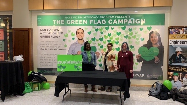 Three people at a booth posing in front of a display case containing a large Green Flag Campaign poster in the IMU
